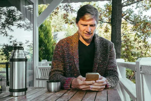 Young man in knitted cardigan drinking coffee on cozy wooden balcony and checking social media by mobile phone. Autumn vibe.