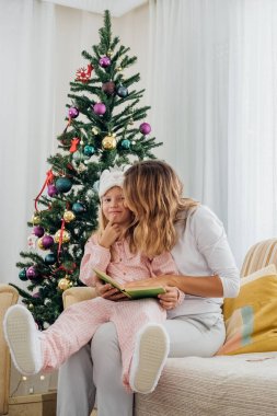 Evdeki Noel ağacının yanındaki kanepede kış masalları anlatan bir kitap okurken küçük kızına sarılan bir kadın..