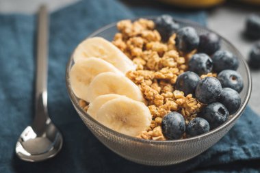 Granola cereal oatmeal with blueberries and banana fruits in a bowl on a blue napkin and grey background.
