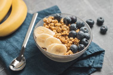 Granola cereal oatmeal with blueberries and banana fruits in a bowl on a blue napkin and grey background.