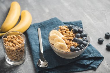 Granola cereal oatmeal with blueberries and banana fruits in a bowl on a blue napkin and grey background.