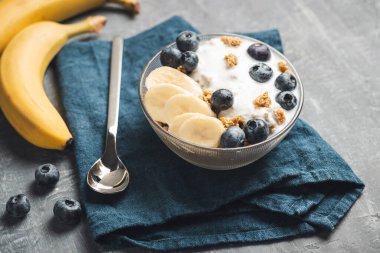 Granola cereal oatmeal with white yogurt, blueberries and banana fruits in a bowl on a blue napkin, grey background.