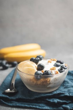 Granola cereal oatmeal with white yogurt, blueberries and banana fruits in a bowl on a blue napkin, grey background.