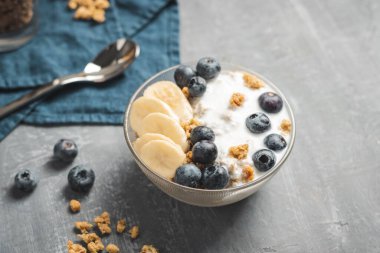 Granola cereal oatmeal with white yogurt, blueberries and banana fruits in a bowl on a blue napkin, grey background.