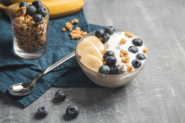 Granola cereal oatmeal with white yogurt, blueberries and banana fruits in a bowl on a blue napkin, grey background.