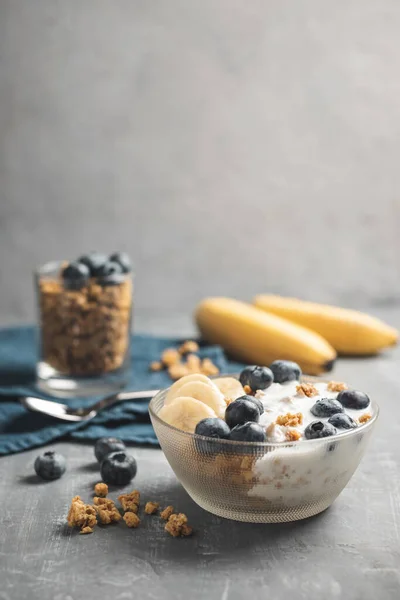 Granola cereal oatmeal with white yogurt, blueberries and banana fruits in a bowl on a blue napkin, grey background.