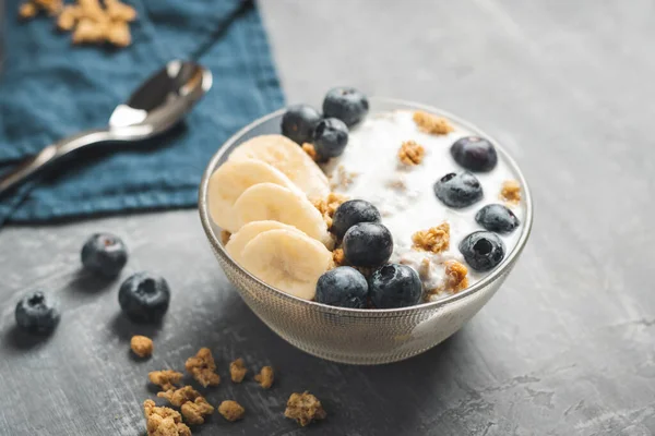 Granola cereal oatmeal with white yogurt, blueberries and banana fruits in a bowl on a blue napkin, grey background.
