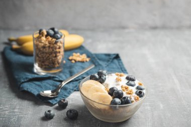 Granola cereal oatmeal with white yogurt, blueberries and banana fruits in a bowl on a blue napkin, grey background.