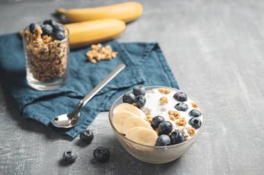 Granola cereal oatmeal with white yogurt, blueberries and banana fruits in a bowl on a blue napkin, grey background.