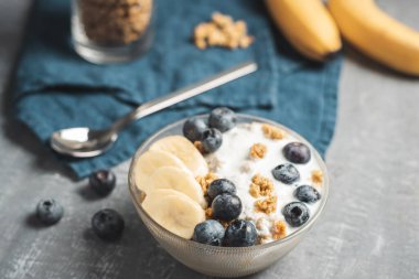 Granola cereal oatmeal with white yogurt, blueberries and banana fruits in a bowl on a blue napkin, grey background.