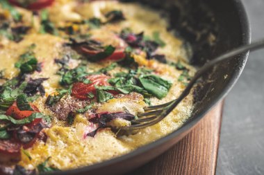 Cooked omelette with sausage, cheese, tomato and herbs in a pan, on wooden board, close-up view.