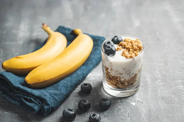 Granola cereal oatmeal with white yogurt, blueberries and banana fruits in a glass on a blue napkin, grey background.