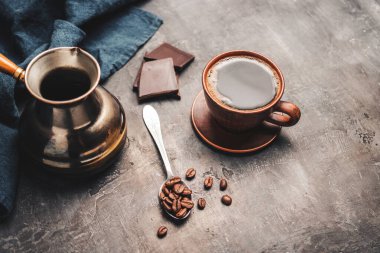 Kil fincanda sade kahve, Türk jezve kahve demliği, çikolata parçaları ve koyu arkaplanda kaşık kahve çekirdekleri..
