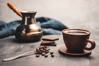 Kil fincanda sade kahve, Türk jezve kahve demliği, çikolata parçaları ve koyu arkaplanda kaşık kahve çekirdekleri..