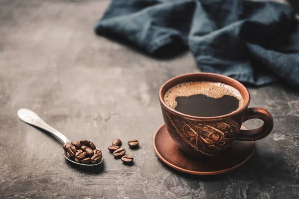 stock image Black coffee drink in a brown clay cup and coffee beans in a spoon on dark background.