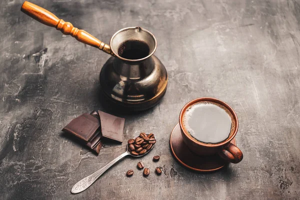 stock image Black coffee drink in a clay cup, turkish jezve coffee pot, chocolate pieces and coffee beans in a spoon on dark background.