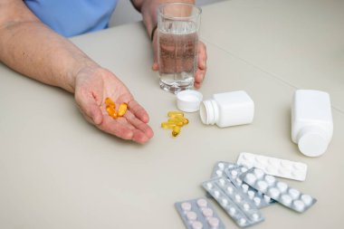 Omega 3 sarı kapsülleri, balık yağı hapları ve su bardağını tutan yaşlı ve buruşuk elleri olan yaşlı bir kadın. Sağlık ve Tıp Konsepti.