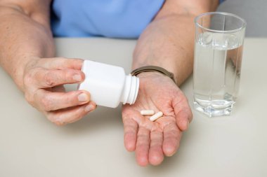 Masada oturan yaşlı kadın tedavi için beyaz kapsüllerden, vitaminlerden ya da haplardan eski kırışık el, sağlık ve ilaç konseptine döküyor..