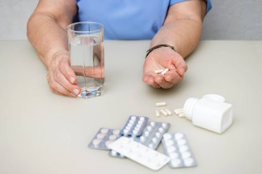 Masada oturan yaşlı kadın, kırışık ellerinde beyaz kapsüller, vitaminler ve tedavi için haplar, su bardağı, sağlık ve ilaç konseptini tutuyor..