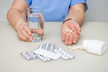 Masada oturan yaşlı kadın, kırışık ellerinde beyaz kapsüller, vitaminler ve tedavi için haplar, su bardağı, sağlık ve ilaç konseptini tutuyor..