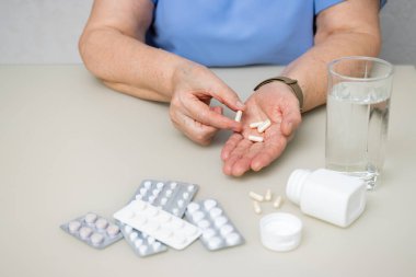 Masada oturan yaşlı kadın kırışık elleriyle beyaz kapsüller, vitaminler veya tedavi için haplar, sağlık ve ilaç konseptini tutuyor..
