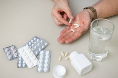 Masada oturan yaşlı kadın kırışık elleriyle beyaz kapsüller, vitaminler veya tedavi için haplar, sağlık ve ilaç konseptini tutuyor..