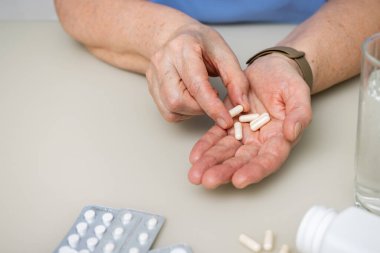 Masada oturan yaşlı kadın kırışık elleriyle beyaz kapsüller, vitaminler veya tedavi için haplar, sağlık ve ilaç konseptini tutuyor..