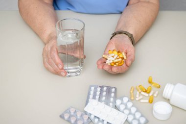Masada kırışık yaşlı elleri olan yaşlı bir kadın. Elinde çeşitli kapsüller ve vitaminler var. Tedavi için avuç içi ilaçlarla dolu. Sağlık ve Tıp Konsepti.