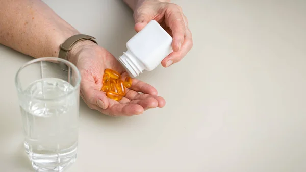 Masada kırışık yaşlı elleri olan yaşlı bir kadın. Omega 3 şişesinden avuç içine sarı kapsüller döküyor. Sağlık ve Tıp Konsepti.