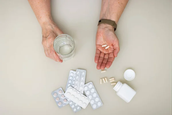 stock image Senior woman sitting at the table and holding in the old wrinkled hands white capsules, vitamins or pills for treatment and water glass, healthcare and medicine concept, top view.