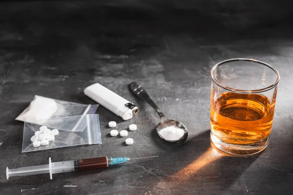 stock image Alcohol drink in a glass, syringe with a dose of drugs, white pills in a transparent bag and narcotics powder in a spoon on dark background. Concept of addiction and bad habits.
