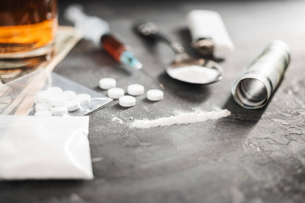 Alcohol drink in a glass, syringe with a dose of drugs, white pills in a transparent bag, narcotics powder in a spoon and US dollar cash on dark background. Concept of addiction and bad habits.