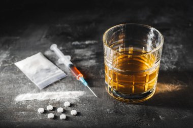 Bardakta alkol içkisi, üzerinde bir doz uyuşturucu, beyaz haplar ve siyah arka planda şeffaf bir torbada uyuşturucu tozu bulunan şırınga. Bağımlılık, istismar ve kötü alışkanlıklar kavramı.