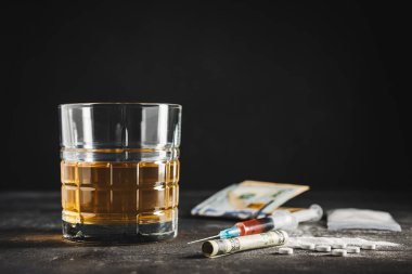 Bardakta alkol, içinde bir doz uyuşturucu, beyaz haplar, şeffaf bir çantada uyuşturucu tozu ve karanlık bir arka planda Amerikan doları nakit para bulunan şırınga. Bağımlılık, istismar ve kötü alışkanlıklar kavramı.