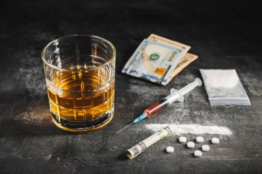Bardakta alkol, içinde bir doz uyuşturucu, beyaz haplar, şeffaf bir çantada uyuşturucu tozu ve karanlık bir arka planda Amerikan doları nakit para bulunan şırınga. Bağımlılık, istismar ve kötü alışkanlıklar kavramı.