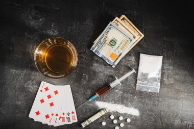 Bardakta alkol içmesi, poker oynamak için kağıt oynaması, bir doz uyuşturucu, beyaz haplar, barut narkotiği ve siyah arka planda Amerikan doları parası olan şırınga. Bağımlılık ve kumar kavramı. en üstteki görünüm.