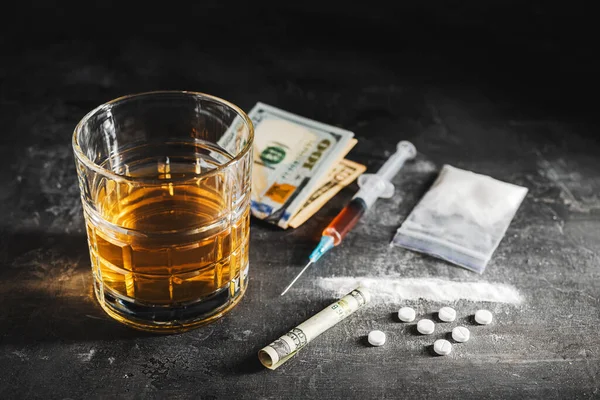 Bardakta alkol, içinde bir doz uyuşturucu, beyaz haplar, şeffaf bir çantada uyuşturucu tozu ve karanlık bir arka planda Amerikan doları nakit para bulunan şırınga. Bağımlılık, istismar ve kötü alışkanlıklar kavramı.