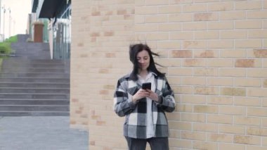 Güzel gülüşü olan çekici genç bir kadın şehrin caddesinde akıllı telefon kullanıyor, rüzgarlı bir bahar günü.