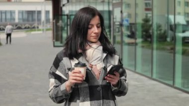 Çekici ve hoş bir genç kadın internet ve sosyal medya için akıllı telefonu kullanıyor, kağıt bardaktan kahve içiyor ve rüzgarlı bir bahar gününde şehir caddesinde yürüyor.
