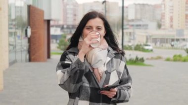 Çekici ve hoş bir genç kadın internet ve sosyal medya için akıllı telefonu kullanıyor, kağıt bardaktan kahve içiyor ve rüzgarlı bir bahar gününde şehir caddesinde yürüyor.
