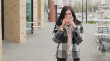Çekici ve cana yakın genç bir kadın gülümsüyor ve şehirdeki bir sokakta kağıt bardaktan kahve içiyor.