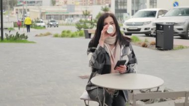 Çekici ve hoş bir genç kadın bir sokak kafeteryasında masada oturuyor, kağıt bardaktan kahve içiyor ve internet ve sosyal medya için akıllı telefonu kullanıyor.