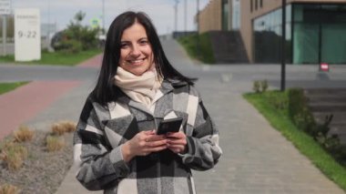 Çekici ve hoş bir genç kadın gülümsüyor, caddede yürüyor ve internet ve sosyal medya için akıllı telefon kullanıyor, yavaş çekim.