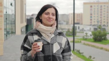 Çekici ve hoş bir genç kadın gülümsüyor, kağıt bardaktan kahve içiyor ve şehirde ağır çekimde yürüyor.