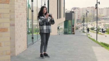 Çekici ve hoş bir genç kadın şehrin bir caddesinde eski model bir kamerayla fotoğraf çekiyor.