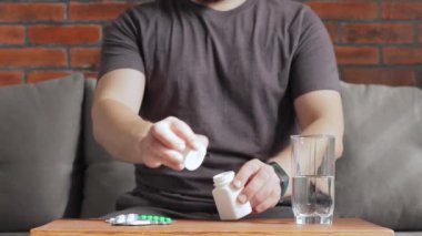 Adam kanepede oturmuş ilaç ya da vitamin hapı alıyor ve bardaktan su içiyor.
