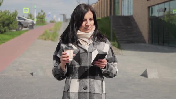 インターネットの閲覧やソーシャルメディアにスマートフォンを使用して魅力的で快適な若い女性 紙コップからコーヒーを飲み 街の通りを歩いて スローモーション — ストック動画