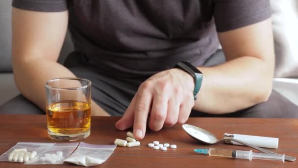 中毒の男はソファに座ってガラスからアルコールを飲んで注射器と白い粉の麻薬でテーブルの上に薬と薬を飲んで — ストック動画