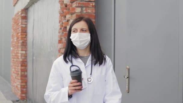病院の裏庭で屋外でコーヒーを飲む聴診器付きの医療用マスクと白いコートの疲れ若い女性医師の肖像 — ストック動画