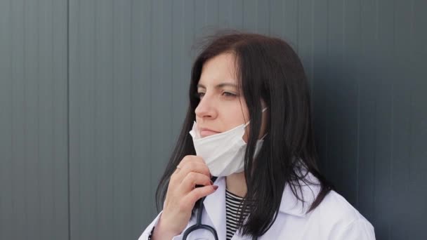 病院の裏庭で屋外でコーヒーを飲む聴診器付きの医療用マスクと白いコートで疲れ若い女性医師の肖像画 スローモーション — ストック動画